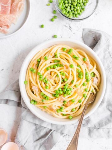 Light Pasta Carbonara with peas and prosciutto on a white plate with eggs in the background